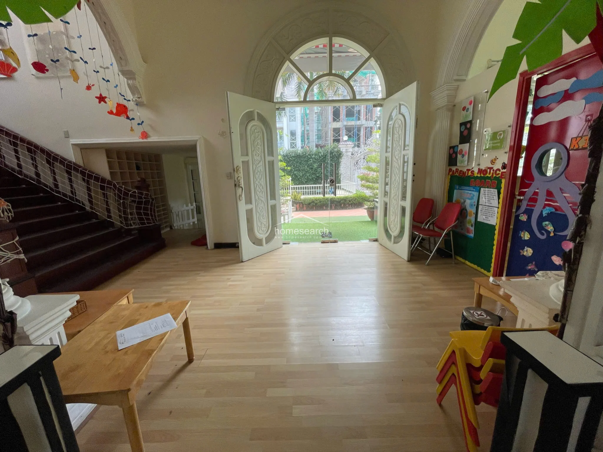 This grand entrance hall features high ceilings with an arched glass doorway that allows for ample natural light and a view of the exterior garden. The space is finished with light wood-look flooring and includes a prominent wooden staircase, currently fitted with safety netting, leading to the upper level.