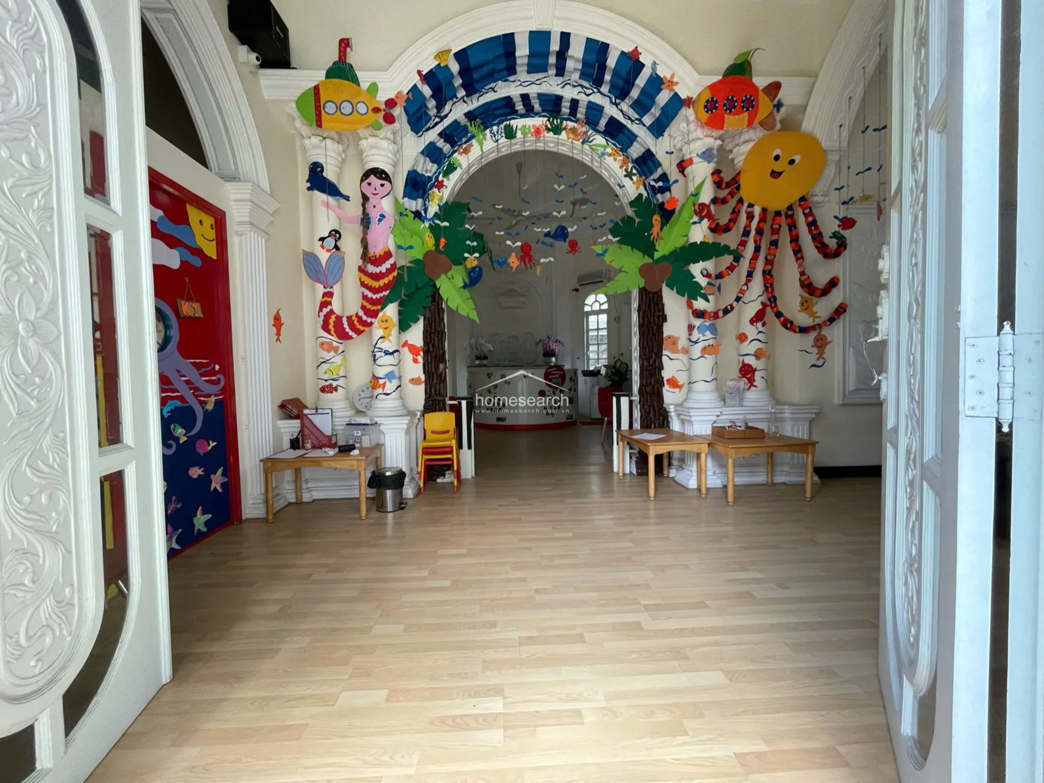This entrance lobby serves as a whimsical, child-friendly reception area characterized by high ceilings, classical columns, and an ornate arched entryway. The space is decorated with vibrant, sea-themed paper crafts and features light-toned wood flooring, creating an inviting atmosphere for a school or daycare setting.