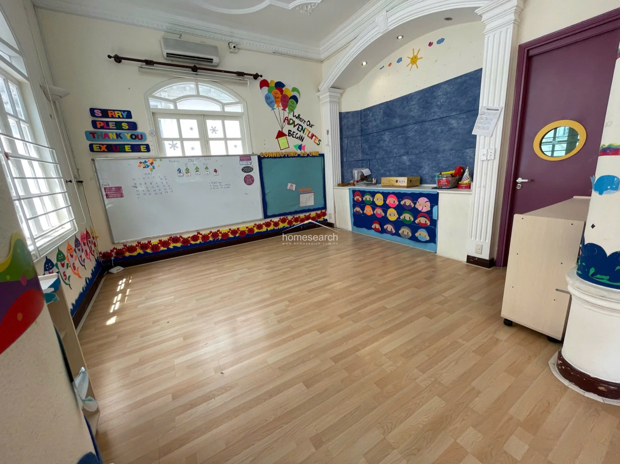 This classroom space features light wood-look flooring and cream-colored walls adorned with educational wall decals and a whiteboard. The room is highlighted by an arched architectural alcove with recessed lighting, crown molding, and a wall-mounted AC unit, creating a bright, functional environment for children.