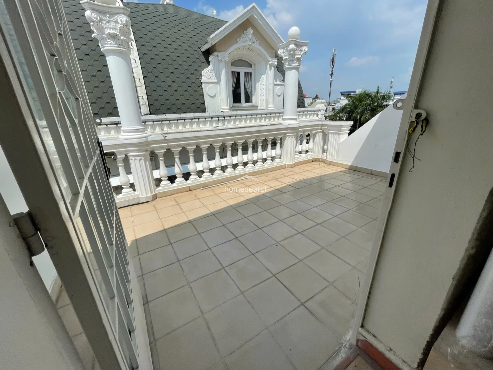This outdoor terrace features a classic neoclassical design with decorative white balustrades and ornate columns, offering an open-air space directly accessible from the interior. The flooring consists of neutral-toned square tiles, and the area provides a clear view of the building's mansard-style roof and architectural detailing. The space appears well-maintained and provides a unique exterior extension to the property.
