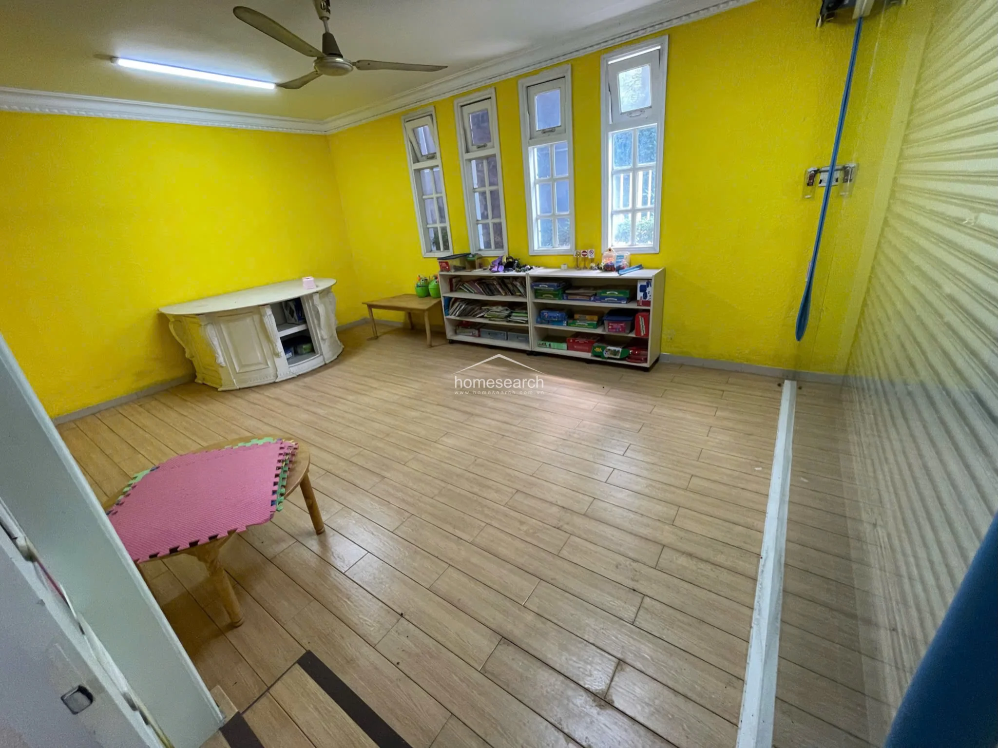 This bright, yellow-walled room serves as a functional play or activity area, featuring four vertical windows that provide consistent natural light. The room is equipped with a ceiling fan, light wood-look flooring, and a low-profile storage shelving unit, maintaining a clean and simple aesthetic.