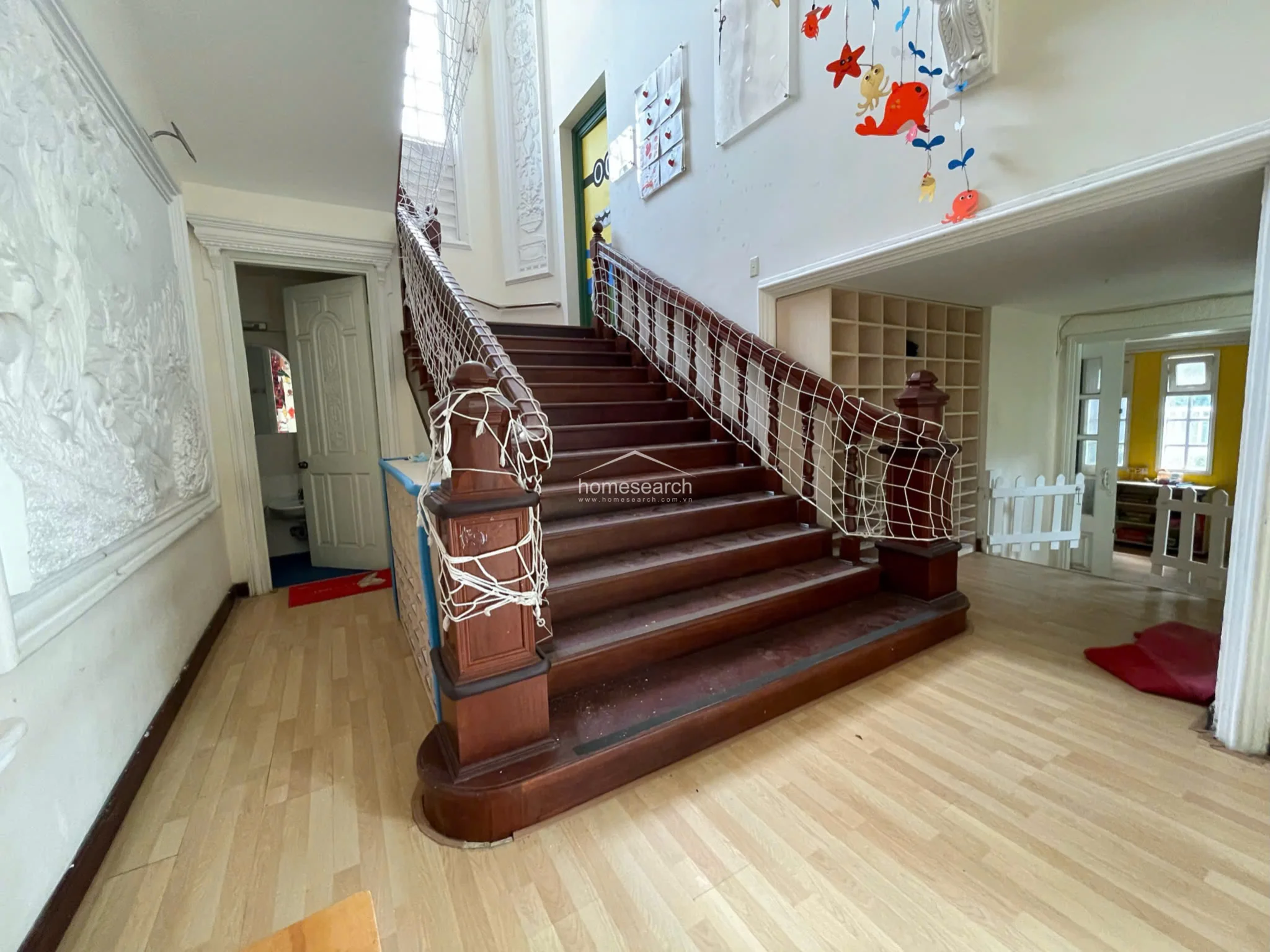 The staircase is a central architectural feature, crafted from rich, dark polished wood and adorned with intricate wall relief carvings. To the side, a large built-in cubby storage unit provides organized space, while the overall condition appears well-maintained but currently repurposed for child-centric use.