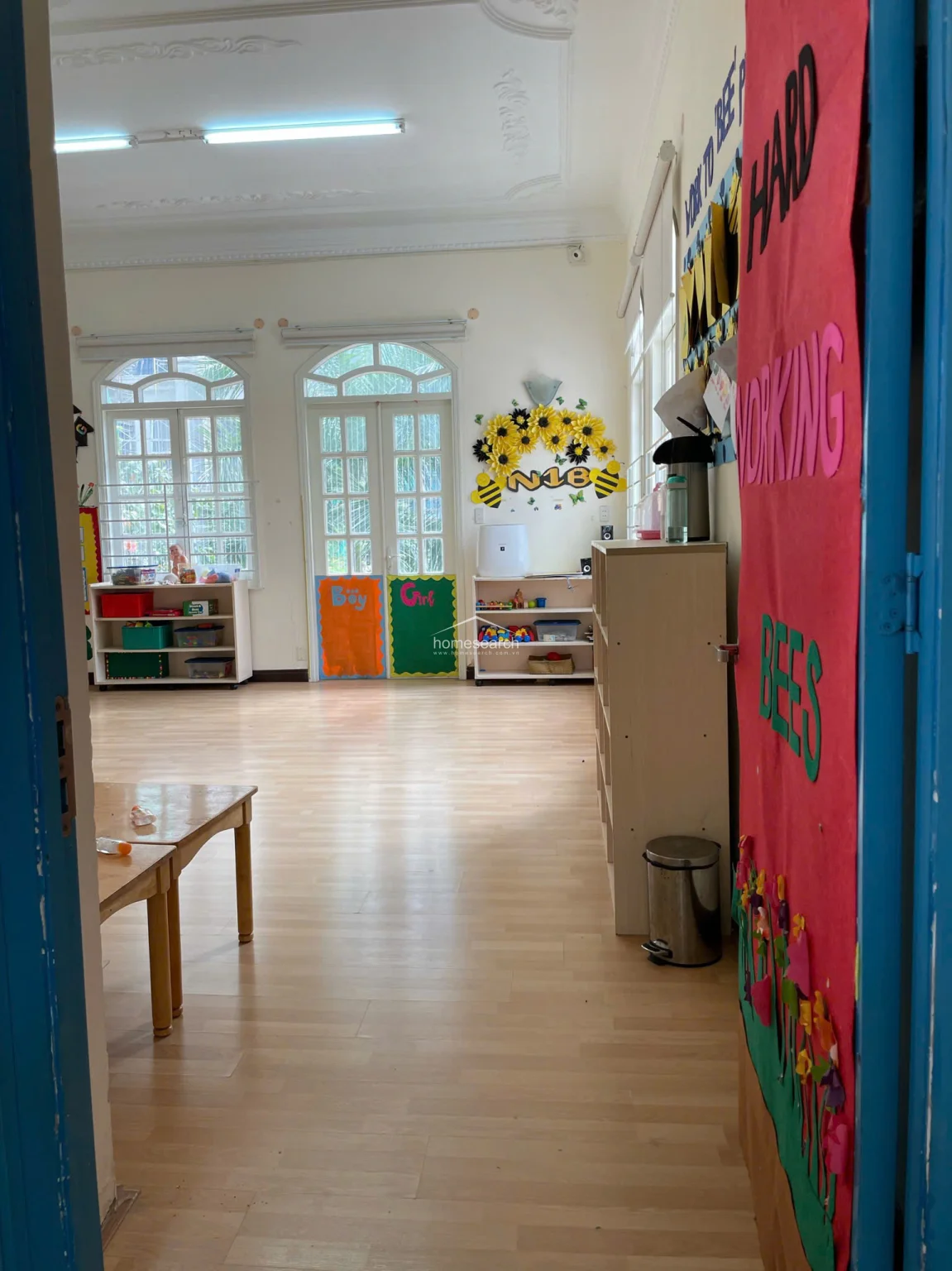 This classroom or activity room features light wood laminate flooring and high ceilings with decorative molding, providing an open and bright environment. The space is organized with low-profile shelving units, educational wall displays, and arched windows that offer views and natural light, maintaining a clean and well-kept appearance.