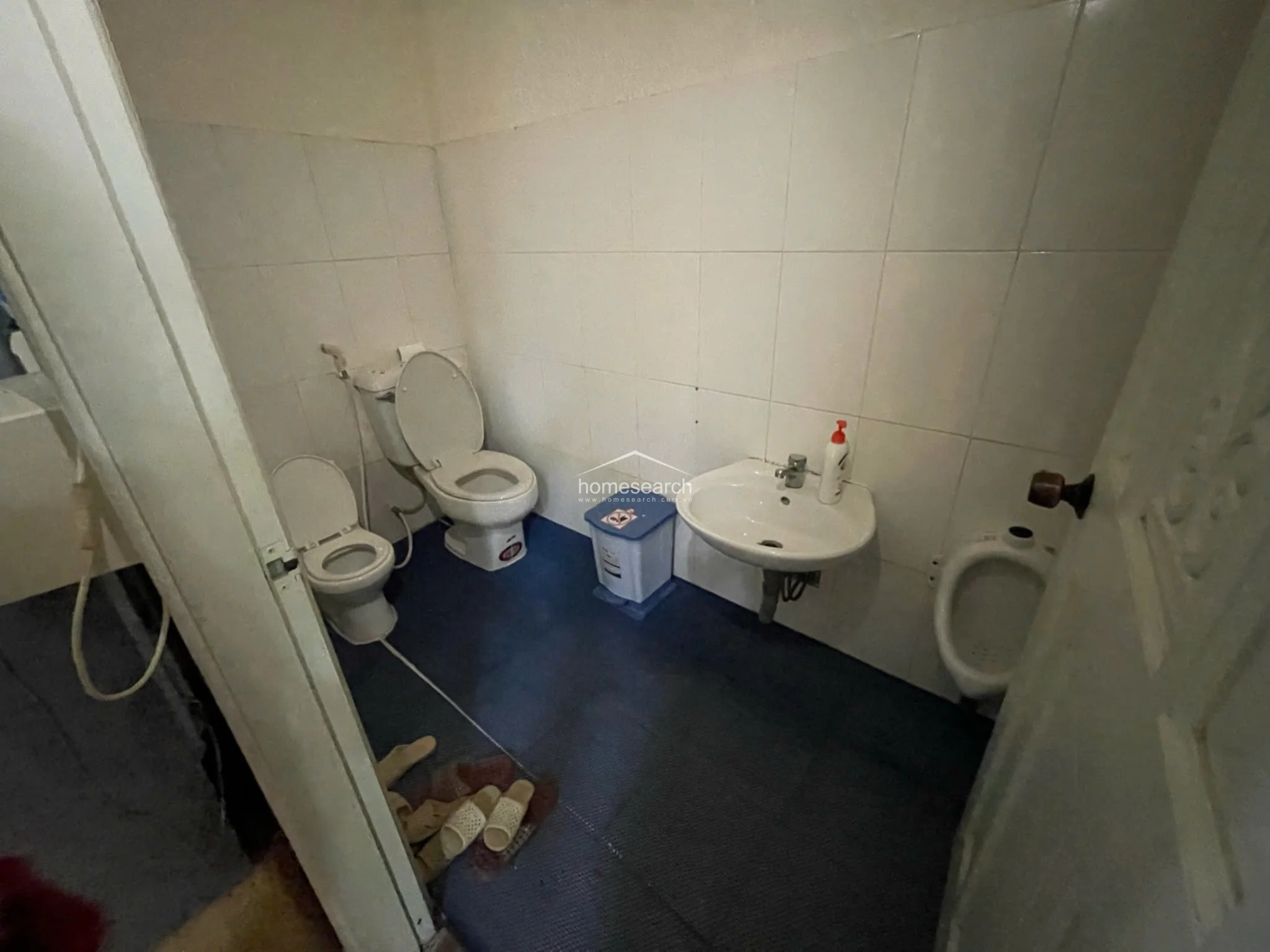 This shared bathroom facility features white wall tiles, a blue non-slip floor covering, and a basic pedestal sink. It is equipped with two toilets and a wall-mounted urinal, indicating a high-traffic commercial or institutional use. The overall condition is utilitarian, with visible signs of wear consistent with its current function.