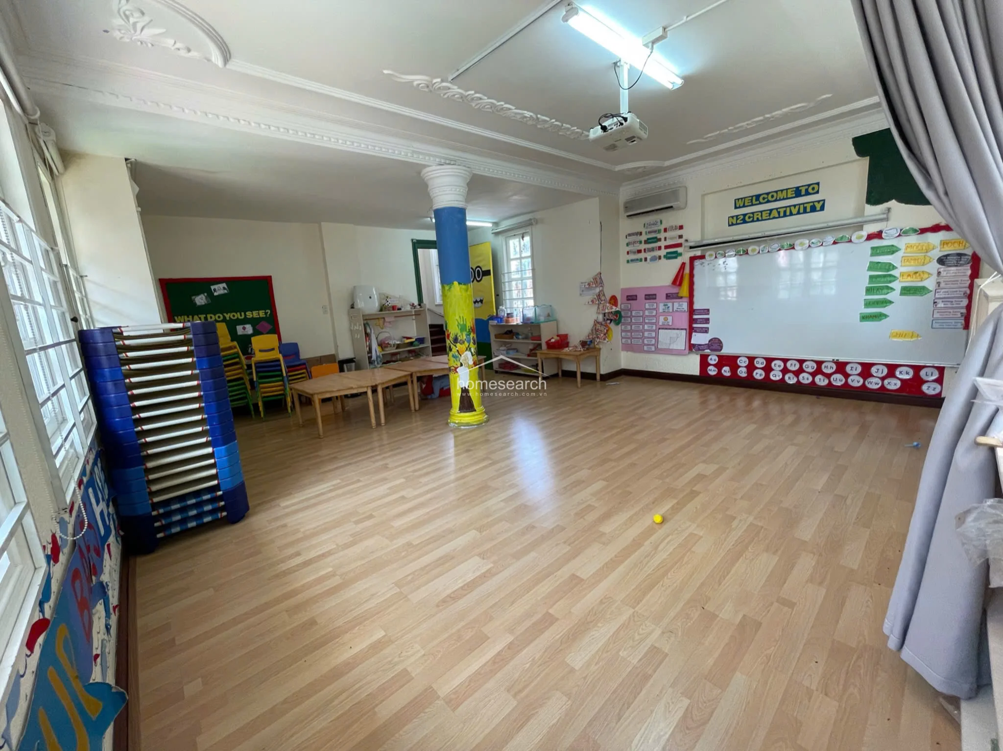 This view of the classroom highlights the functional layout, featuring a large whiteboard and a projector system for interactive learning. The space is well-maintained with light wood flooring and decorative ceiling details, providing a clean and organized environment for students.