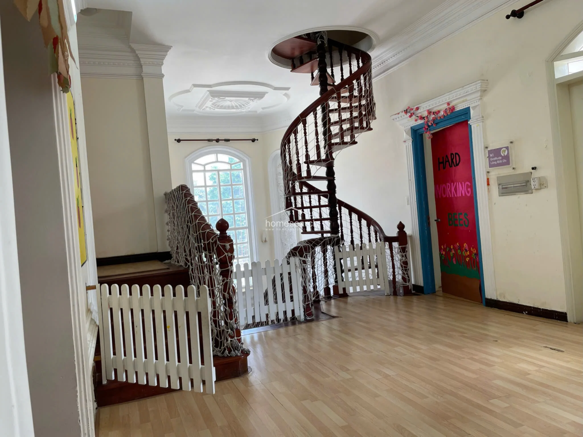 This landing area showcases a striking dark wood spiral staircase as the central architectural feature, complemented by light wood laminate flooring and ornate ceiling molding. The space connects to other rooms through a colorful, themed doorway and features a large arched window that allows for significant natural light.