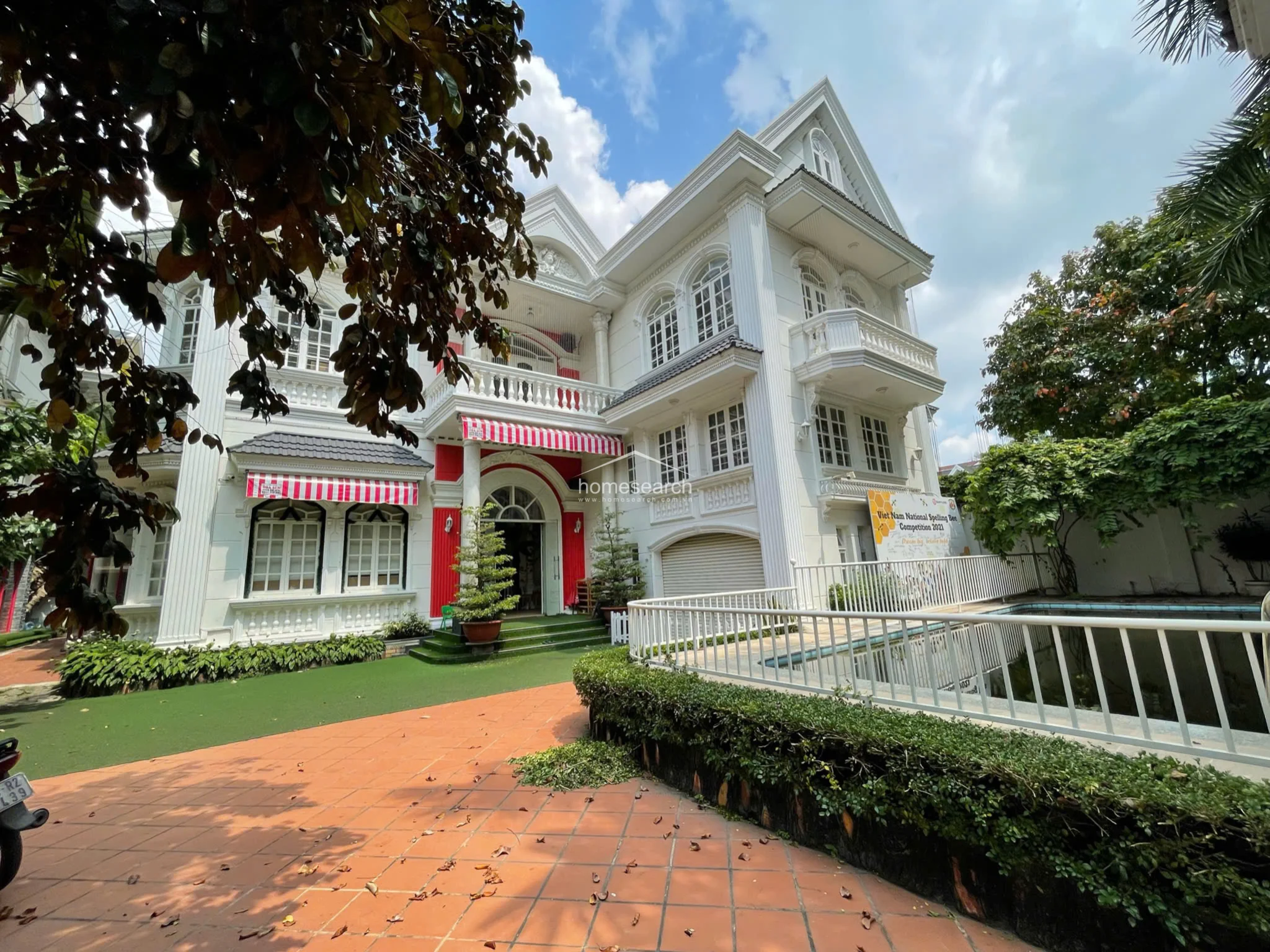 This grand, multi-story neoclassical villa features a striking white facade accented with bold red structural elements and striped awnings. The exterior showcases intricate architectural details, including tall fluted columns, arched windows, and decorative balustrades, set against a backdrop of lush greenery and terracotta-tiled pathways.