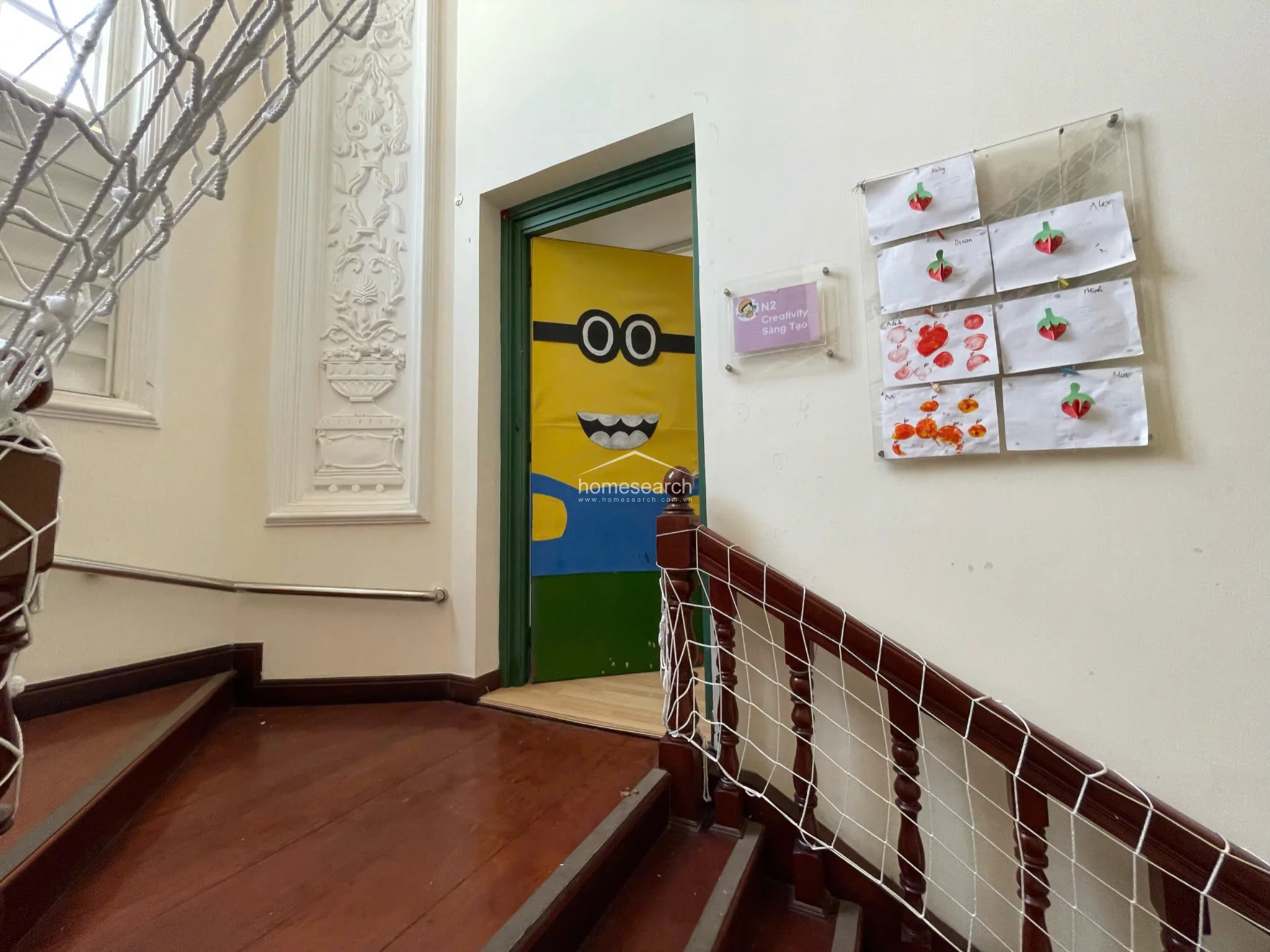 This image shows a staircase landing featuring rich, dark wood flooring and a classic white decorative wall molding. A whimsical Minion-themed door leads into the classroom, while a net-covered wooden banister adds a unique architectural touch.