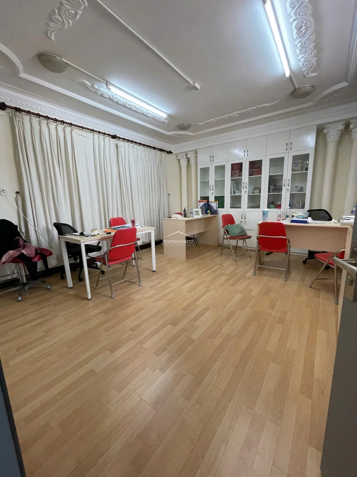 This office or administrative area features elegant white built-in cabinetry with glass-fronted doors, providing ample storage and display space. The room is accented by classic architectural columns and decorative ceiling moldings, creating a professional yet bright atmosphere with light wood flooring and floor-to-ceiling curtains.