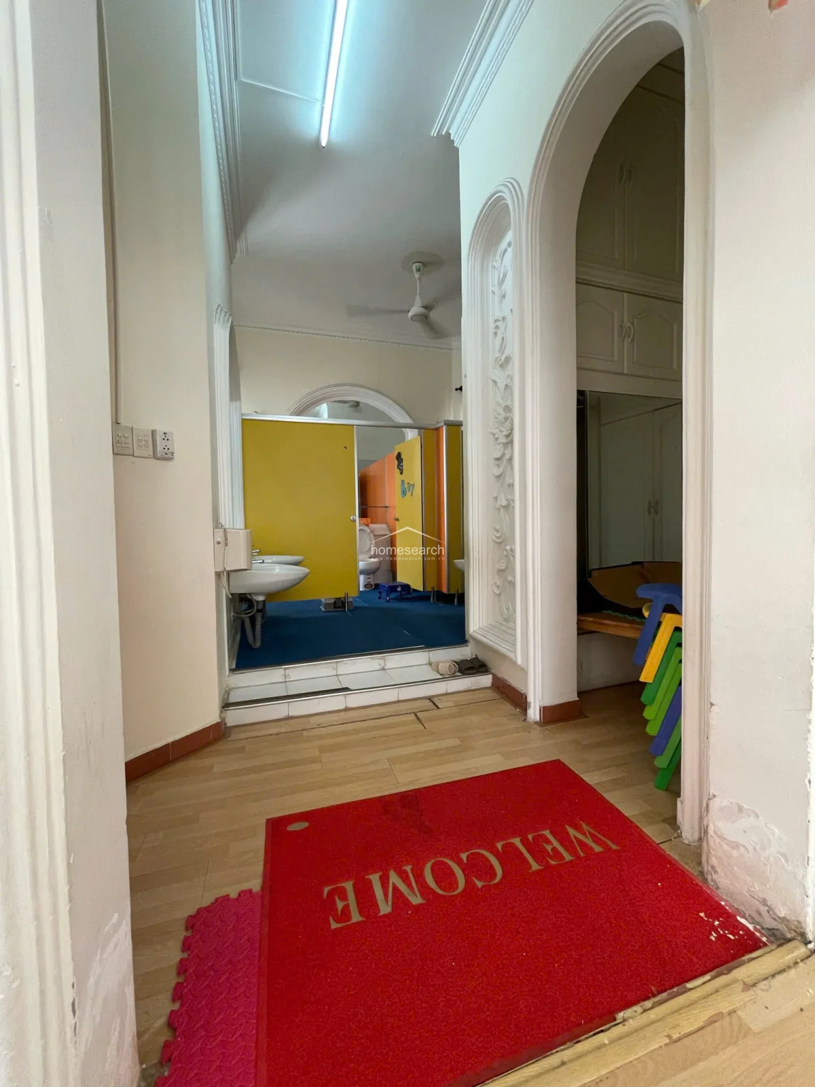 This transitional hallway area features light wood flooring and leads into a restroom facility equipped with vibrant yellow privacy partitions and blue flooring. The space is framed by elegant, ornate white architectural columns and moldings, reflecting a well-maintained, classic interior design style.
