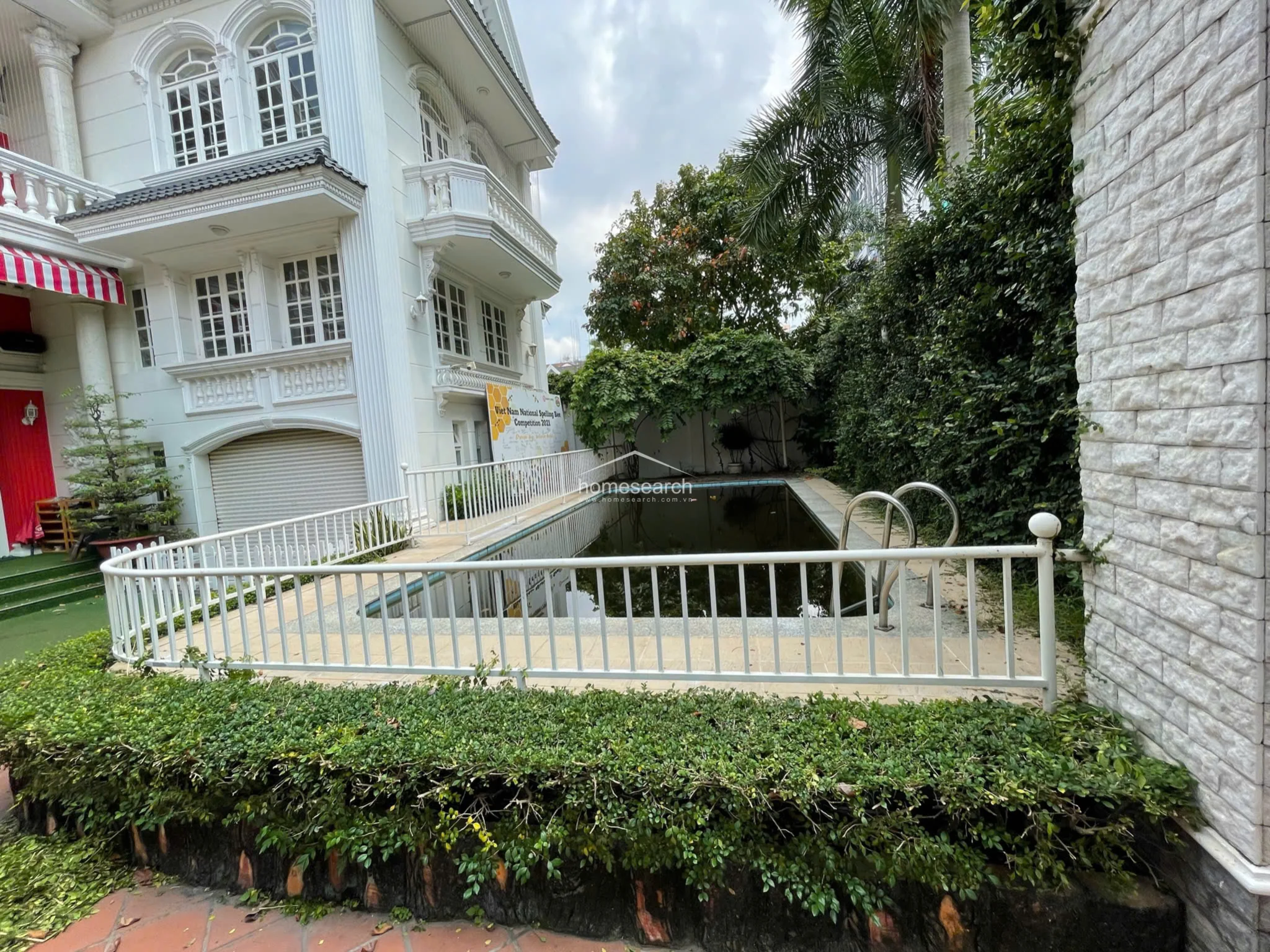 This image highlights the private outdoor pool area, which is enclosed by a white metal safety fence and bordered by dense, mature landscaping. The pool itself appears to be in need of maintenance, as the water is dark and stagnant, contrasting with the well-kept white exterior of the villa visible in the background.