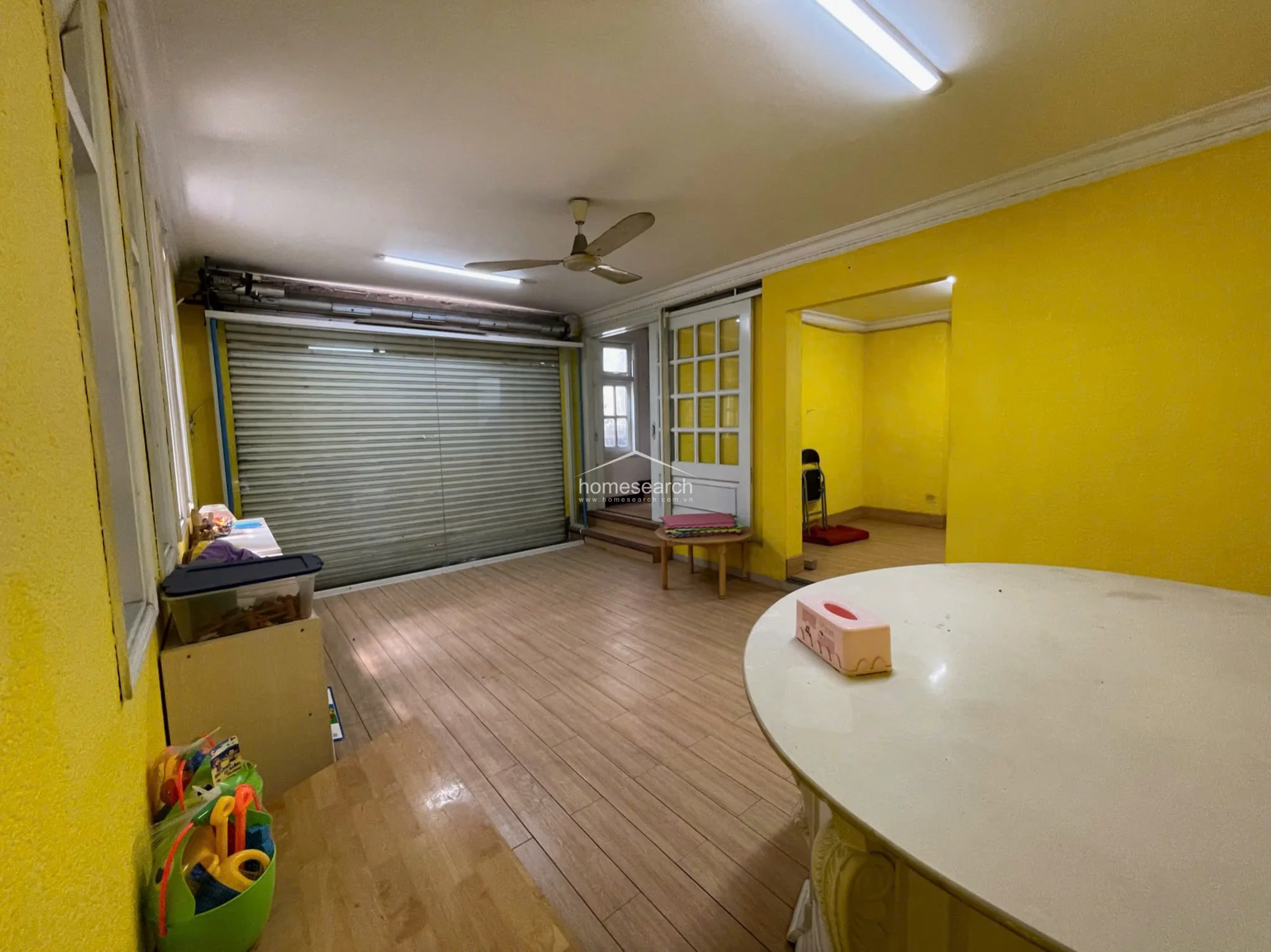 This versatile room features a roll-up metal security shutter, suggesting it may have previously served as a garage or a secure ground-floor workspace. It connects directly to an adjacent room through a white-framed glass door, with bright yellow walls and light wood-look flooring creating a cohesive, airy feel throughout the space.