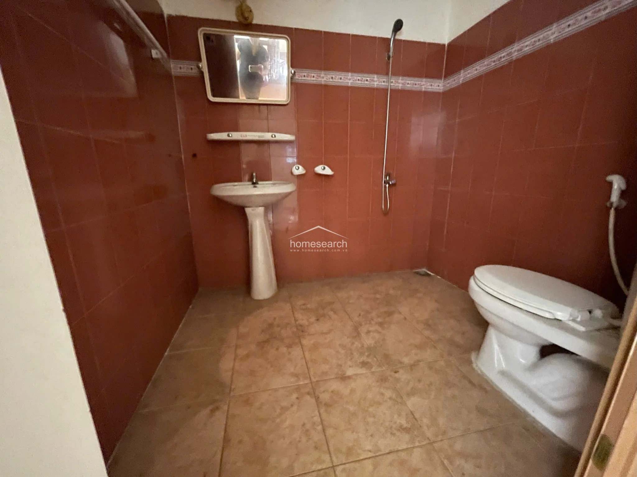 This bathroom features a dated aesthetic with deep red wall tiles and a contrasting beige tiled floor. It is equipped with a standard pedestal sink, a wall-mounted mirror, and a basic shower fixture, reflecting a functional but older condition that may benefit from modernization. The layout is compact and utilitarian, focusing on essential fixtures without additional storage or vanity cabinetry.