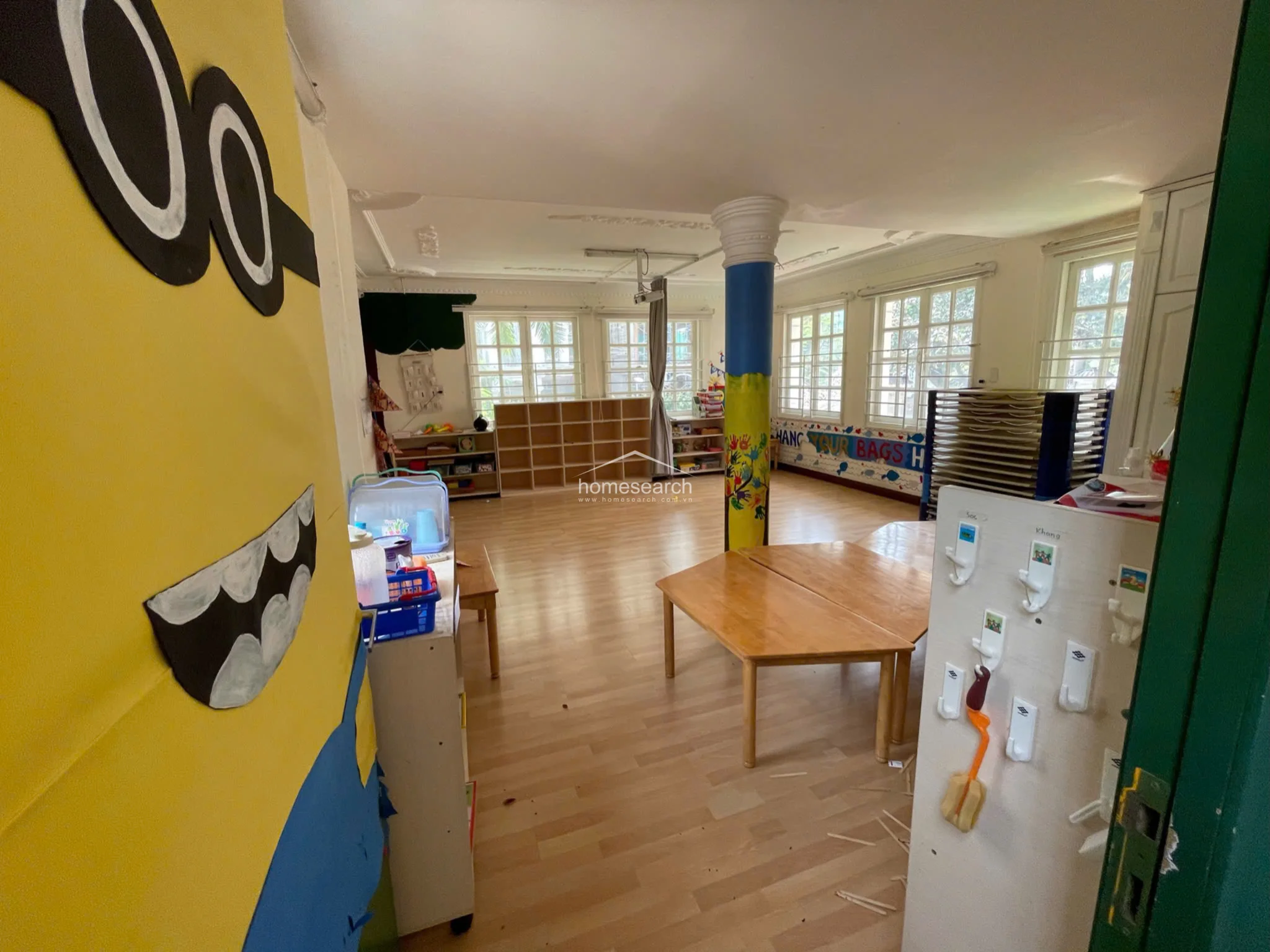 This classroom space features light-toned wood flooring and is anchored by a central structural column painted with a colorful tree mural. Abundant natural light enters through multiple windows, illuminating the open layout which includes modular wooden tables and open shelving units for storage.