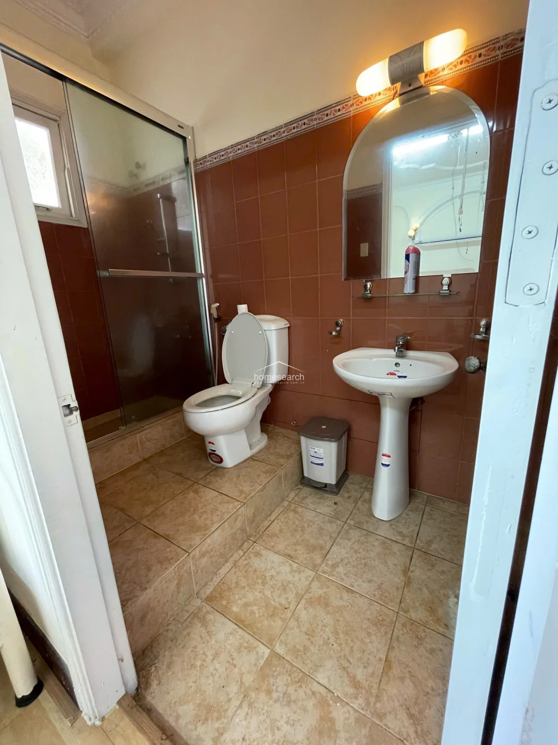 This bathroom features a raised platform for the toilet and a separate glass-enclosed shower stall, finished with dark reddish-brown wall tiles and beige floor tiles. It is equipped with a pedestal sink and a vanity mirror, showing a well-maintained, traditional design.