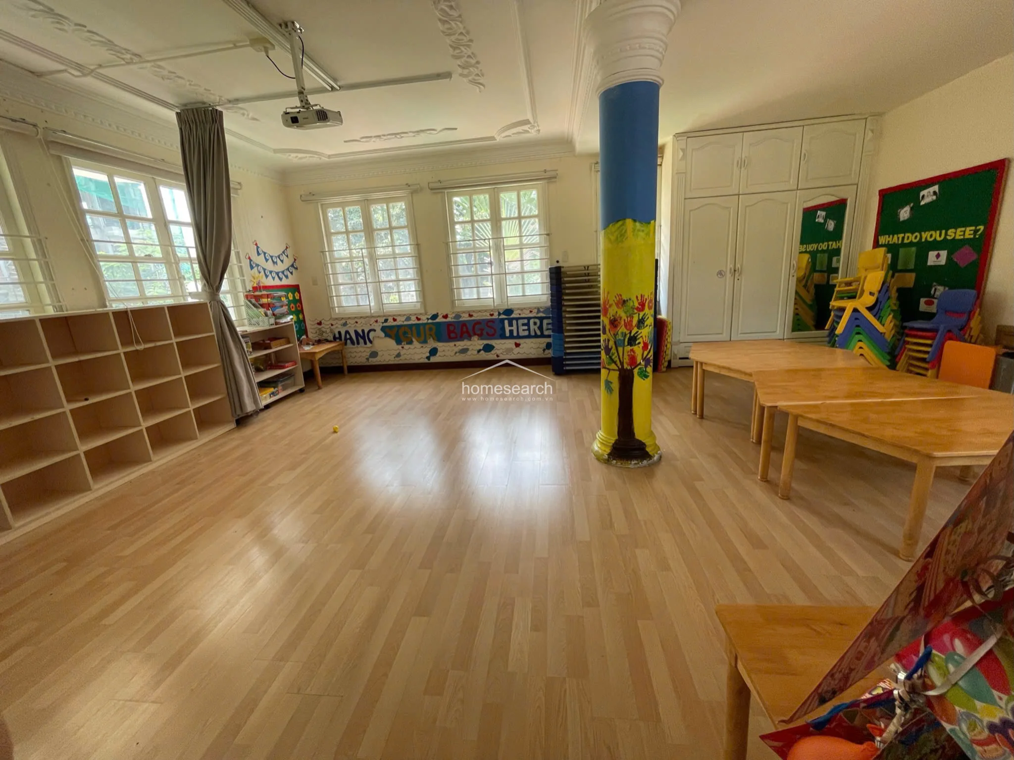 The classroom maintains a bright and airy feel with its light wood flooring and neutral wall palette, complemented by a decorative ceiling cornice. Storage is well-integrated with a large built-in white cabinet unit, while the room is equipped with a ceiling-mounted projector for educational use.