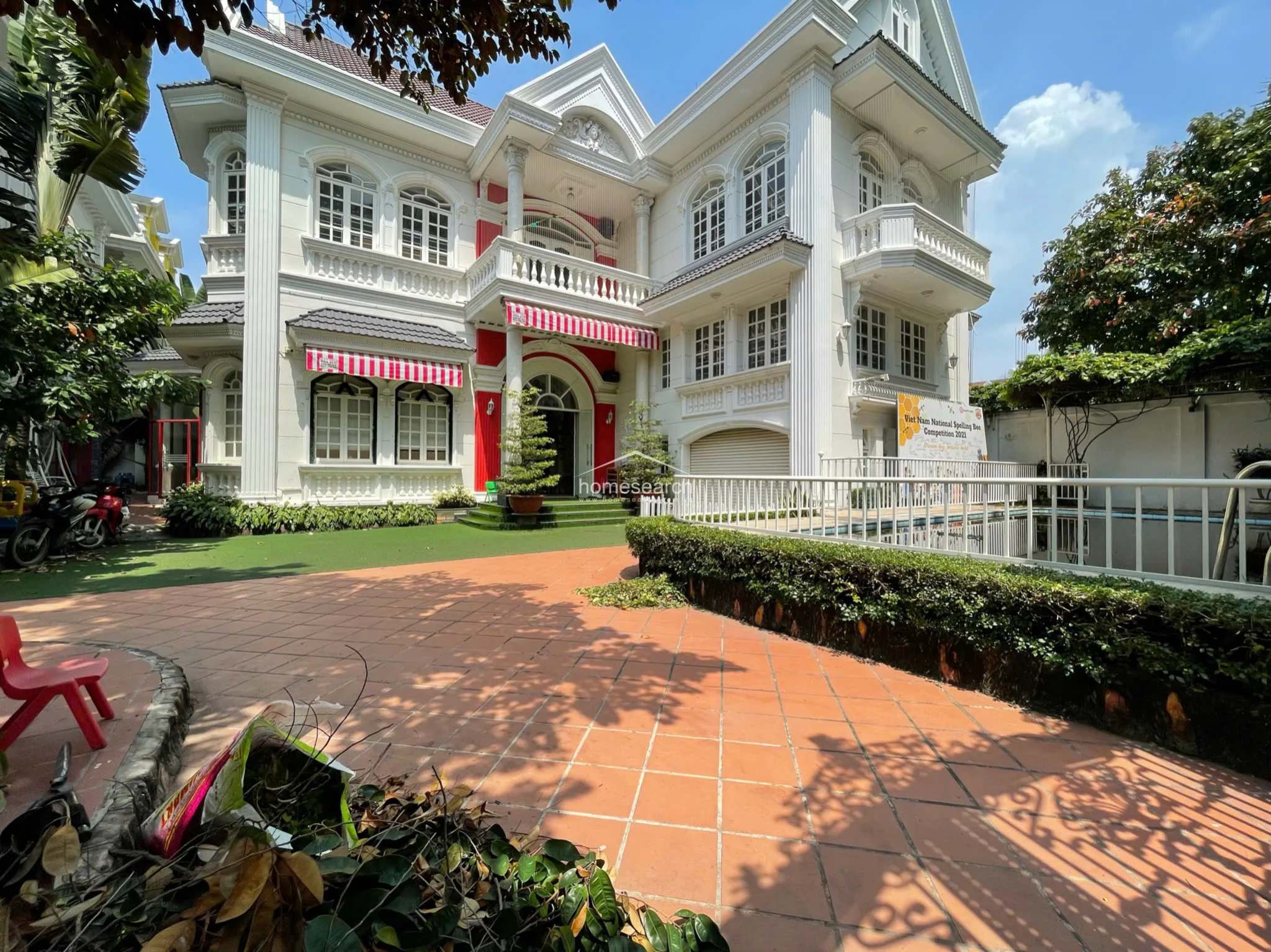 A wide-angle view captures the expansive front courtyard, which transitions from a terracotta-tiled patio to a manicured artificial grass area leading to the main entrance. The property displays a high level of architectural complexity, with multiple balconies and a prominent, ornate arched doorway that serves as the focal point of the facade.