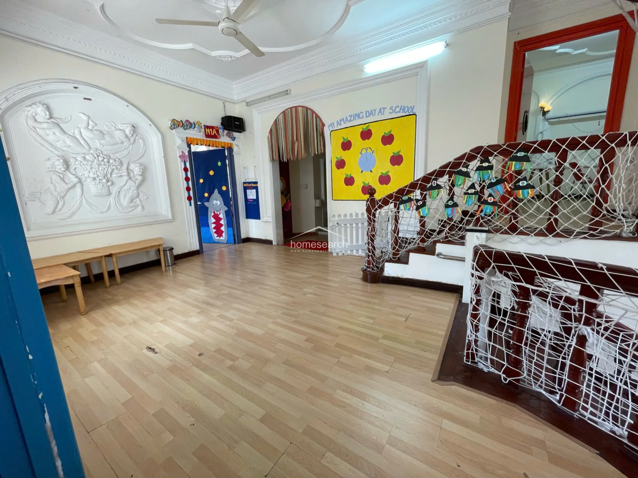 This spacious entrance hall features light-toned wood flooring and an ornate ceiling with decorative molding and a central fan. The space is accented by a classical relief sculpture on the wall and connects to multiple rooms through arched doorways, currently decorated for a school environment.