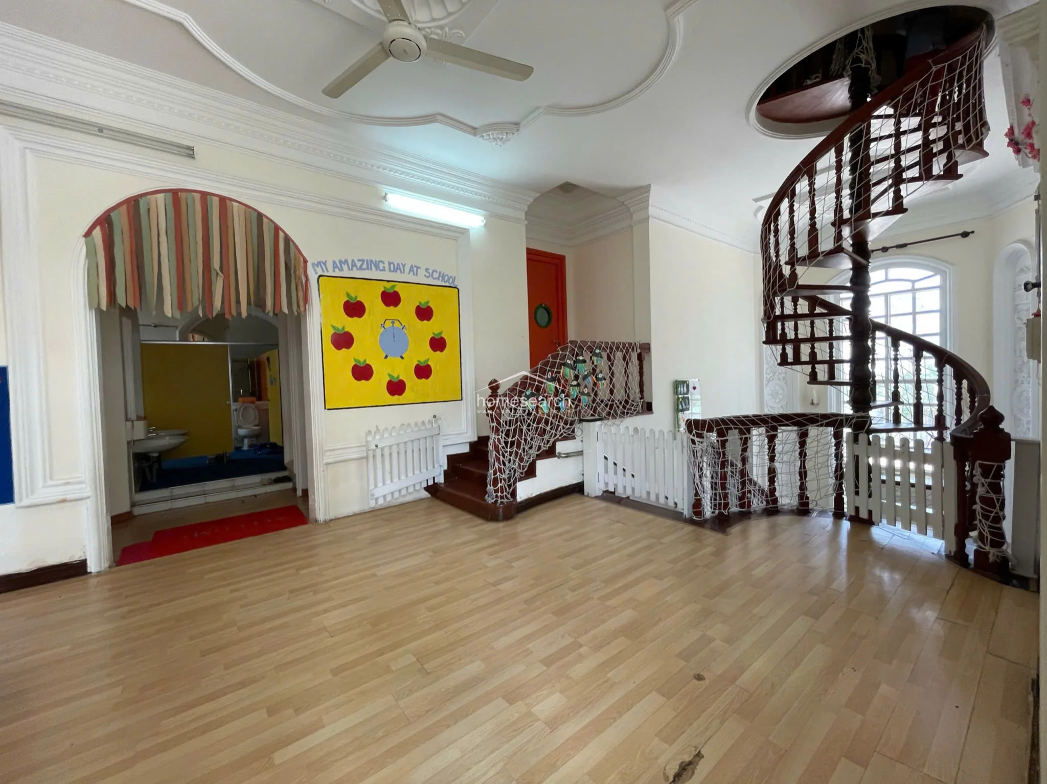 This view highlights the flow between the main hall and an adjacent room, showcasing the grand wooden spiral staircase that serves as a central architectural feature. The area is currently child-proofed with safety netting and white picket-style fencing, reflecting its use as a communal or educational space.