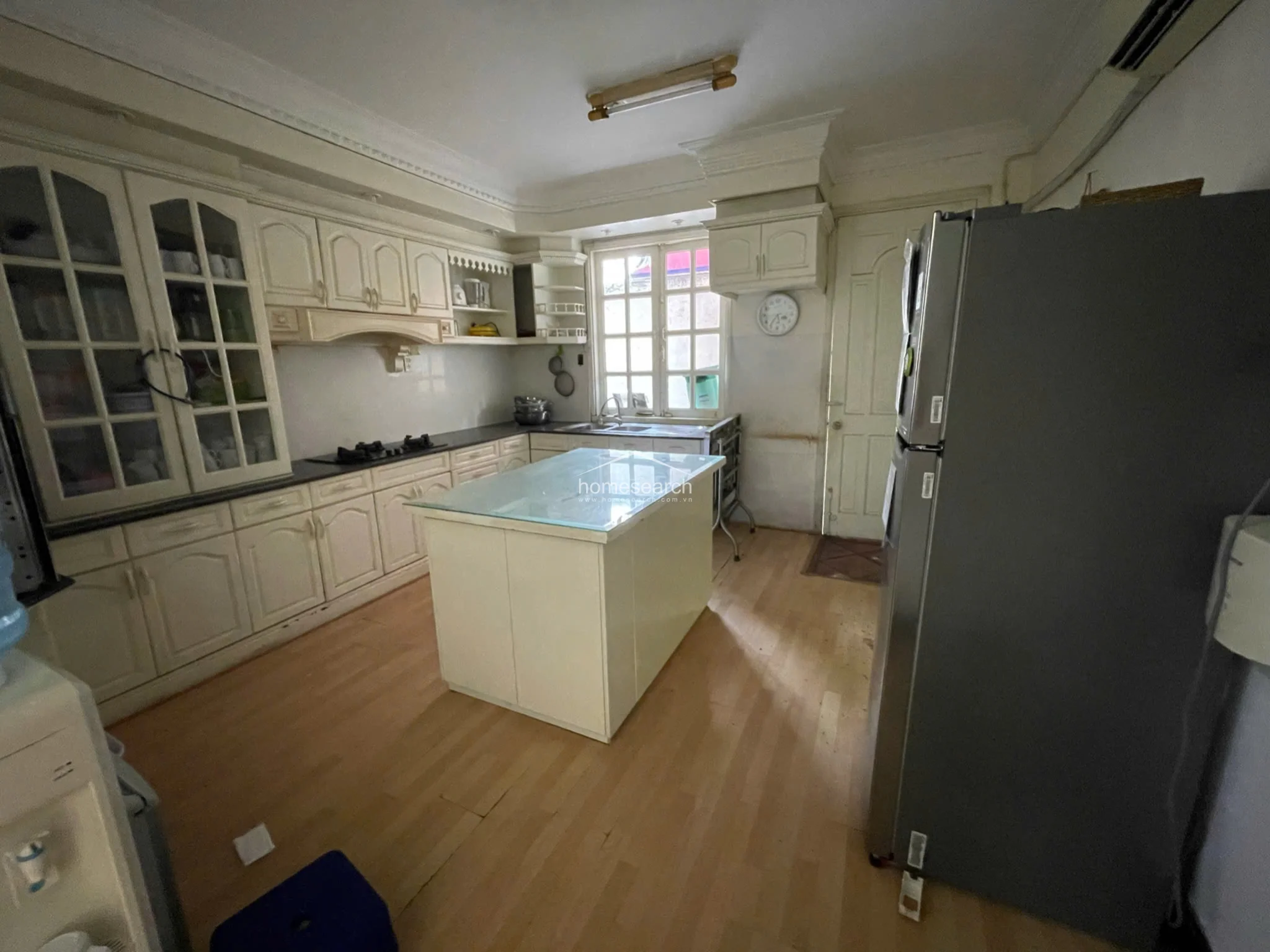 The kitchen is equipped with ample off-white cabinetry, a central island with a glass countertop, and a large stainless steel refrigerator. It features light wood flooring and a classic window that provides natural light, though some minor wall staining suggests a need for cosmetic maintenance. The layout is spacious and functional, offering significant storage space within the built-in cupboards and shelving.