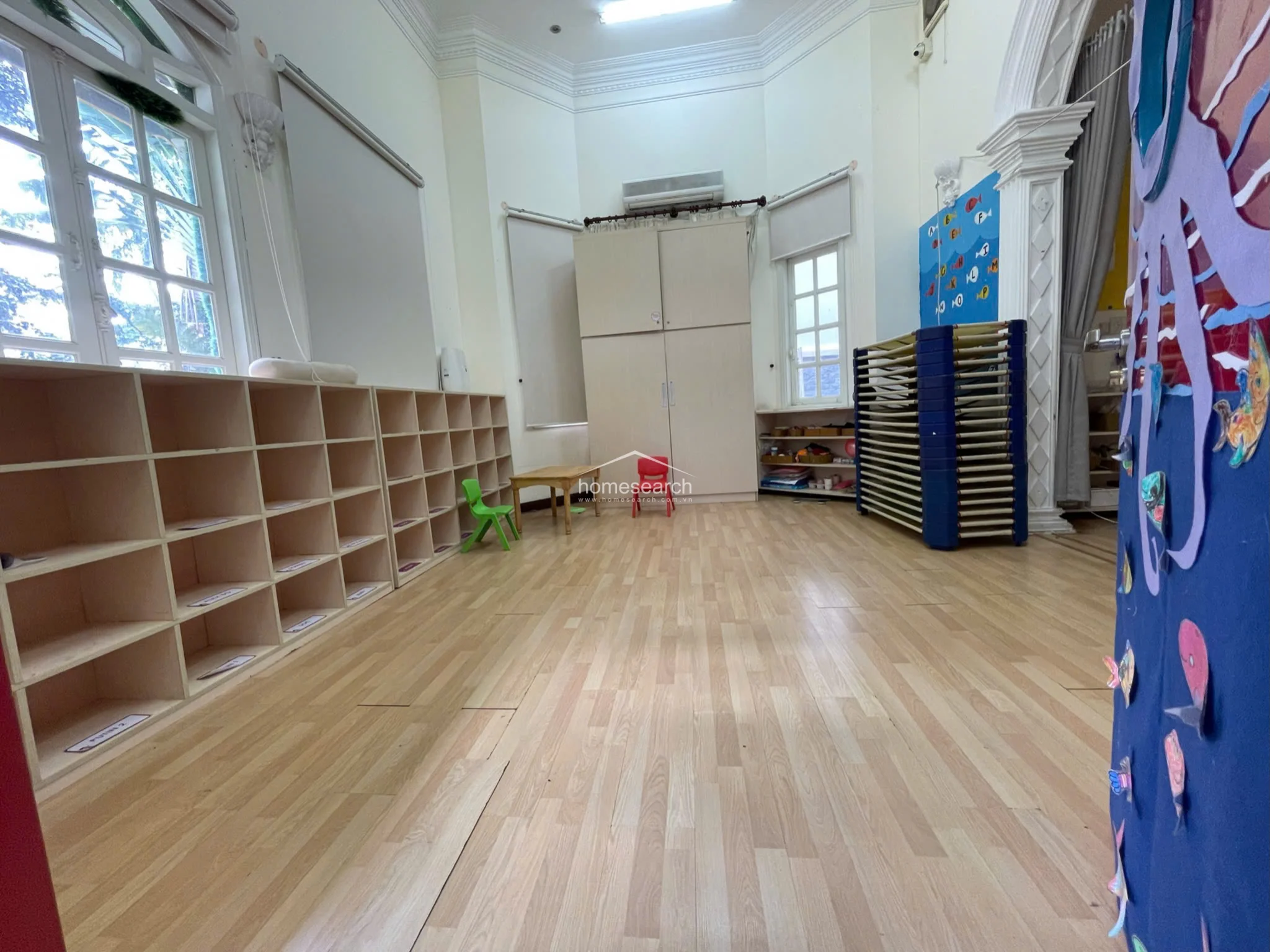 This classroom is designed for organization and functionality, featuring extensive built-in wooden shelving units along the wall for student storage. The room is equipped with a wall-mounted AC unit, light wood flooring, and a stack of foldable cots, providing a clean and structured environment for children.