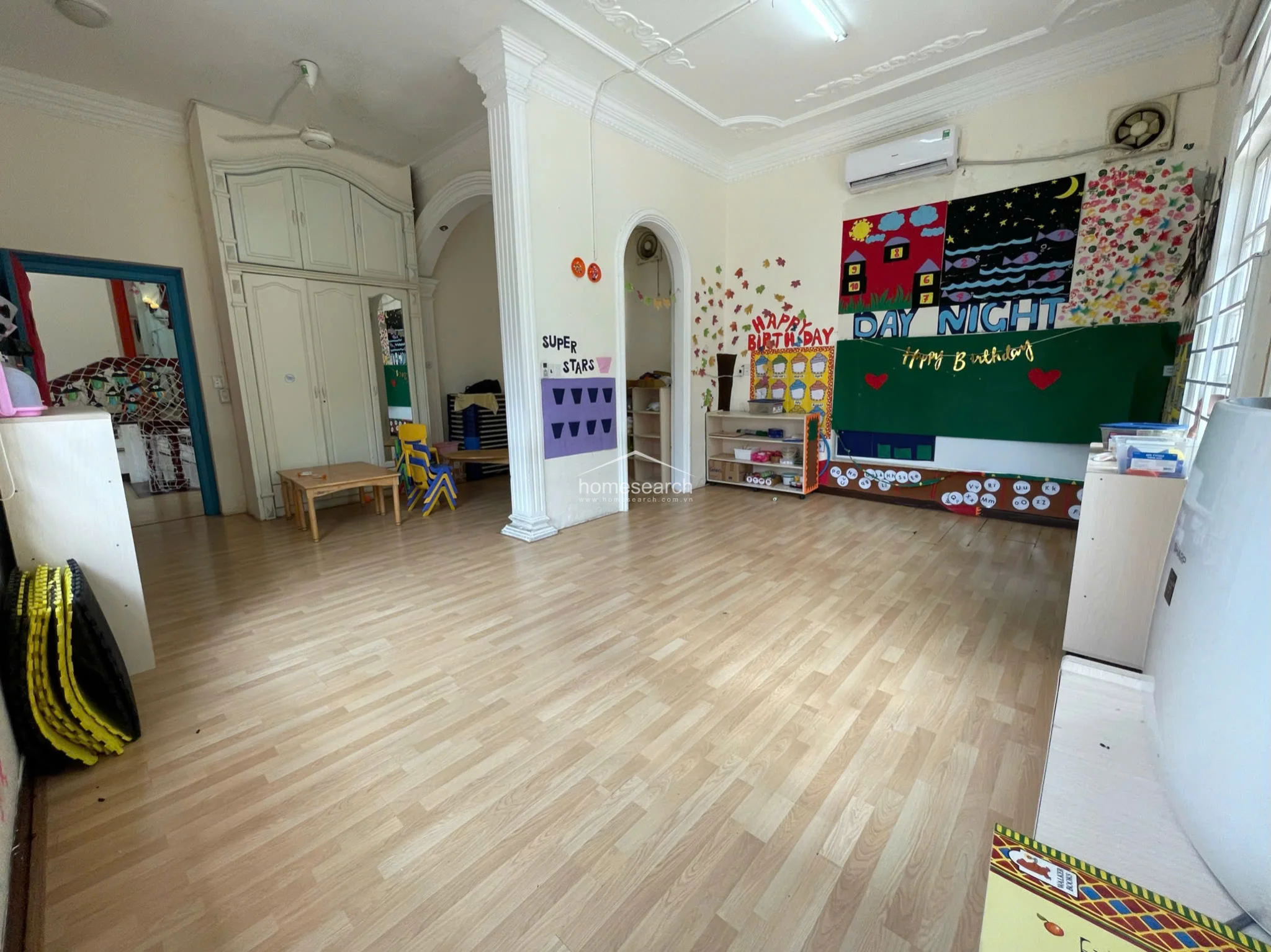 This spacious classroom is characterized by light wood laminate flooring, high ceilings with crown molding, and a prominent white column that divides the room. It includes built-in white cabinetry, a wall-mounted air conditioning unit, and large windows, creating a bright and flexible learning environment with plenty of storage and wall space for educational visuals.