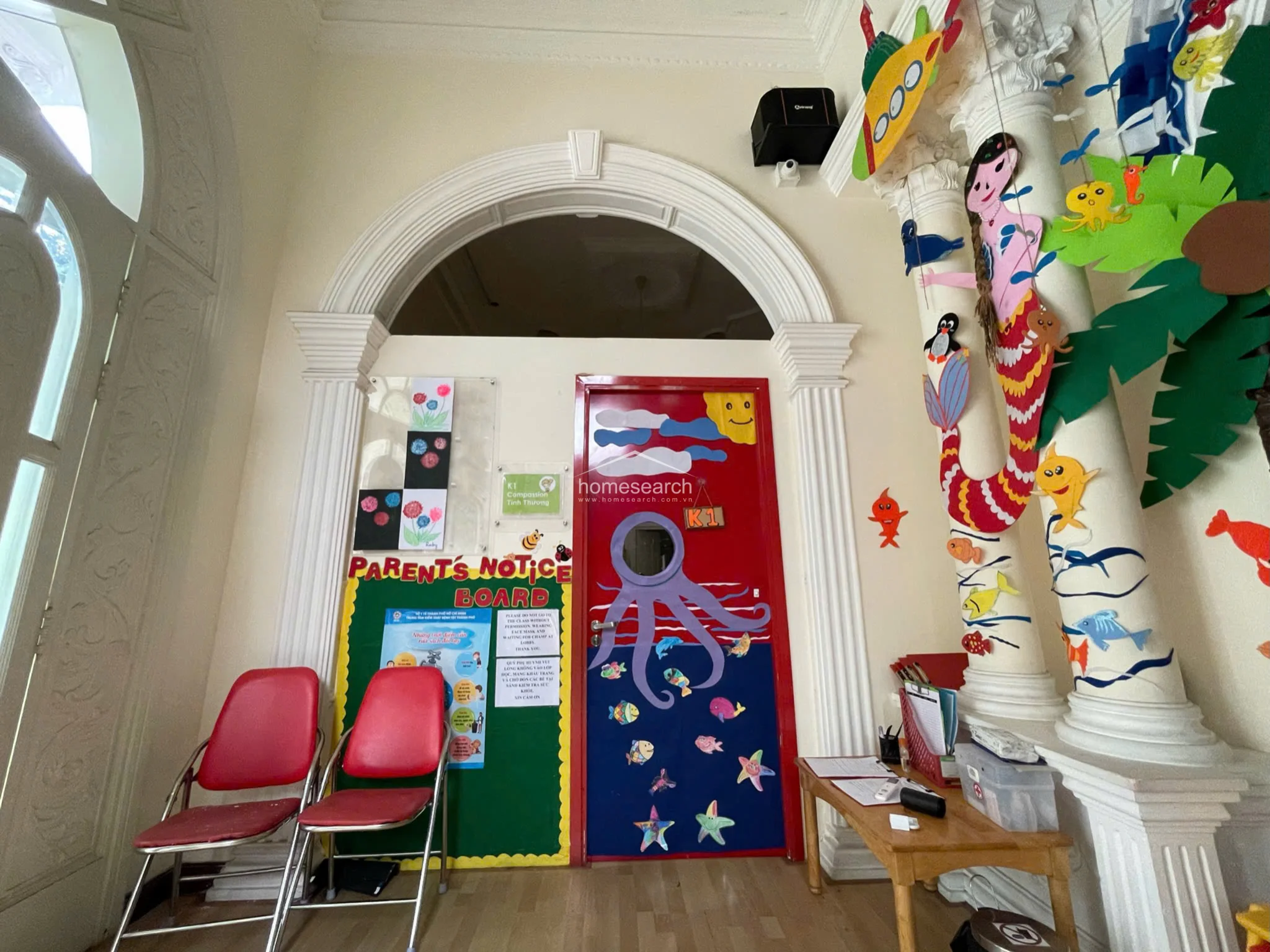 This interior space functions as a classroom or waiting area, featuring a prominent arched doorway and a "Parents' Notice Board" for communication. The room maintains a consistent theme with colorful, ocean-inspired door decorations and child-sized furniture, set against a backdrop of neutral walls and light wood flooring.