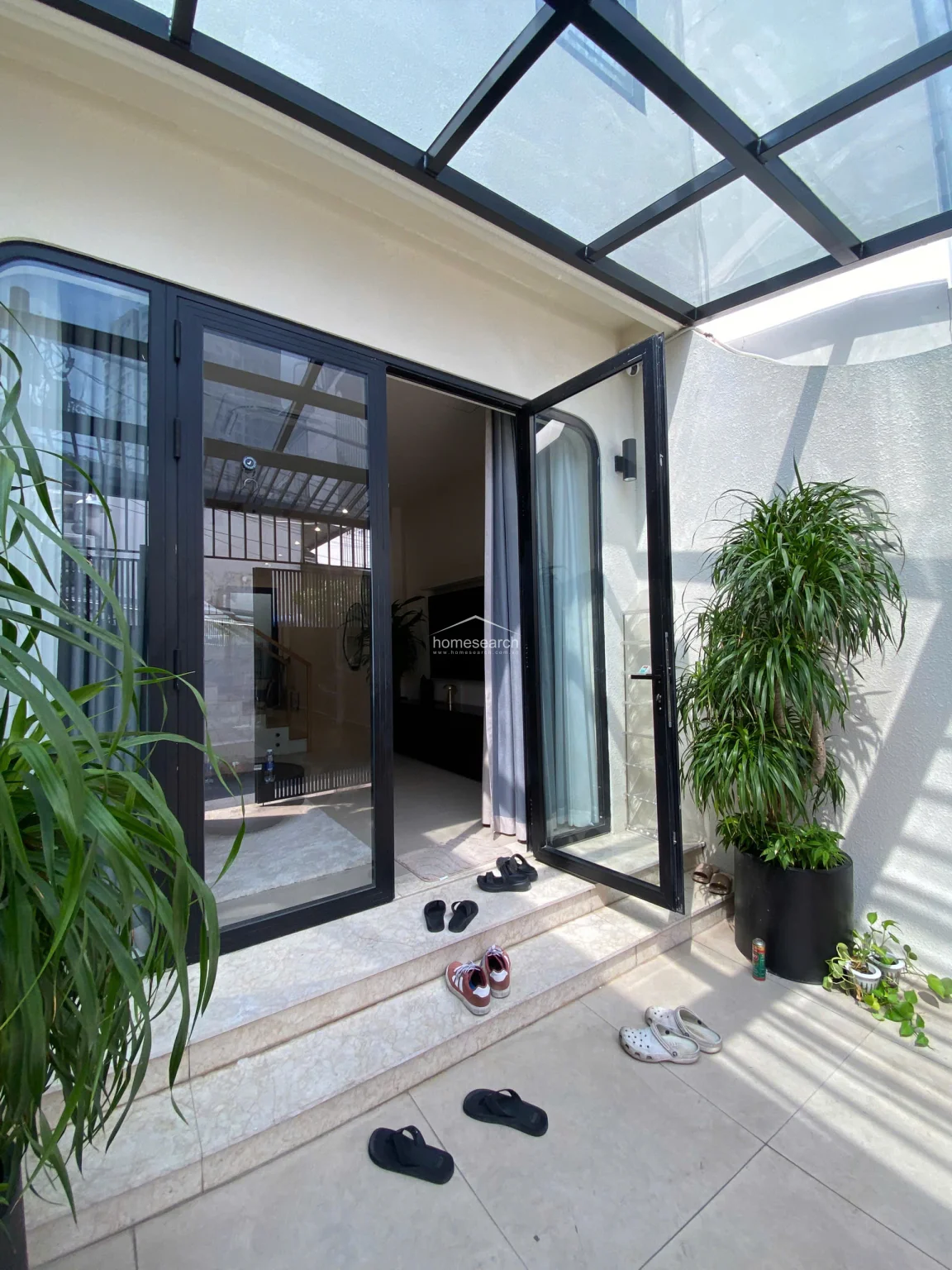 This image captures the transition between the interior living space and a small outdoor terrace, featuring a modern black-framed glass door system. The terrace is partially covered by a glass roof and includes a potted plant, creating a bright, airy connection to the outdoors. The flooring consists of light-colored stone tiles, and the overall condition appears well-maintained and contemporary.