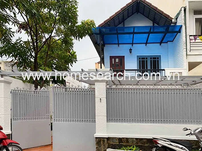 This image shows the exterior of a two-story residential property featuring a light blue facade, a gabled roof, and a private balcony. The property is secured by a grey metal gate and a matching fence, with a translucent blue awning providing overhead shelter for the entrance area.
