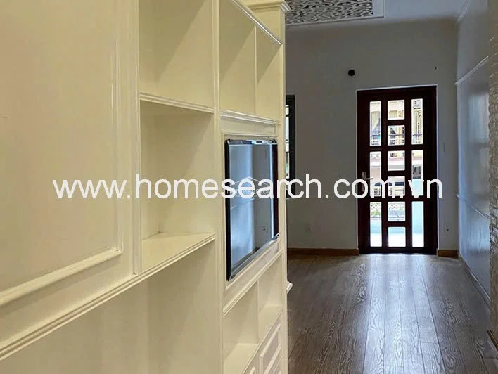 This image displays a living or hallway area featuring a custom-built white shelving unit that integrates a television mount. The space is finished with warm-toned wood flooring and connects to an adjacent room through a dark-framed glass door, which allows for natural light flow. The overall condition appears well-maintained with a clean, modern aesthetic.