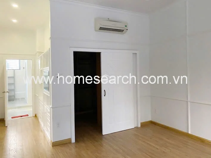 This bedroom or multi-purpose room features a functional layout with a sliding white door that opens into a dedicated walk-in closet or storage area. The room is equipped with a wall-mounted AC unit and is finished with light wood flooring and crisp white walls. The space flows directly into an adjacent tiled area, suggesting a practical and efficient floor plan.