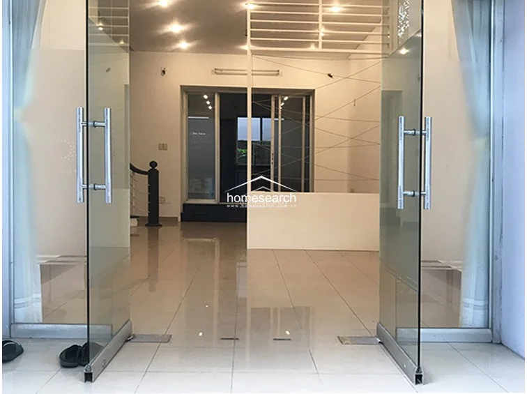 This entrance area features modern glass double doors that open into a spacious, open-plan living area with light-colored tile flooring. The space is bright and airy, leading directly toward a staircase and a rear kitchen area, creating a seamless flow throughout the ground floor.