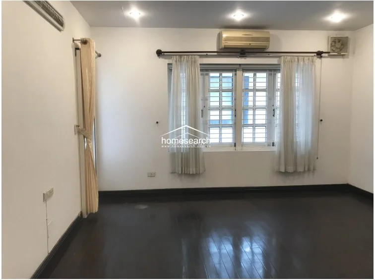 This bedroom or multi-purpose room is characterized by a large, multi-pane window dressed in light-filtering curtains, allowing for ample natural light. The room is equipped with a wall-mounted air conditioning unit and an exhaust fan, while the dark hardwood floors and white walls create a balanced, neutral backdrop.