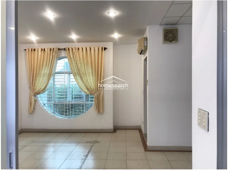 This living area features a distinctive circular window with security bars and elegant beige curtains, allowing for soft natural light. The space is finished with neutral-toned floor tiles and recessed ceiling lighting, creating a clean, minimalist backdrop for furniture placement.