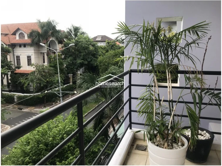 This balcony offers a pleasant view of a quiet, tree-lined residential street and neighboring villas, providing a serene outdoor escape. It is furnished with potted palm plants and features a durable metal railing, making it a well-maintained space for relaxation.