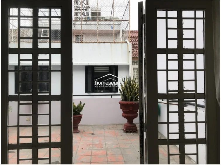 This image shows a view from inside looking out onto a tiled terrace area, framed by white metal security grilles on the doors. The outdoor space features large decorative urns with plants and provides a view of neighboring buildings, suggesting a private urban balcony or rooftop setting.