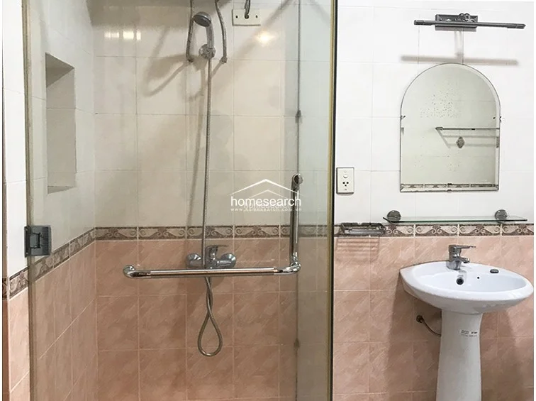 This bathroom is finished in soft pink and white wall tiles with a decorative border, creating a classic aesthetic. It includes a pedestal sink, a glass-enclosed shower with a handheld fixture, and a wall-mounted mirror, maintaining a clean and well-kept appearance.