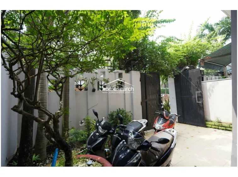 The exterior features a private courtyard or driveway area with mature greenery and a decorative concrete boundary wall. It provides convenient, secure storage for multiple motorbikes and is enclosed by a sturdy metal gate, offering a practical transition between the street and the home.
