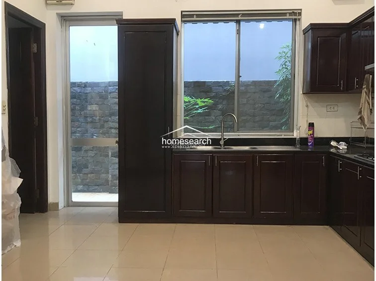 The kitchen is designed with dark wood cabinetry that contrasts sharply against the light tile flooring and white walls, providing ample storage space. It features a large window above the sink that looks out toward a stone-walled outdoor area, ensuring the workspace receives natural light and ventilation.