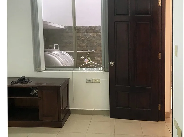 This interior space features light-colored tile flooring and plain white walls, providing a neutral backdrop. A dark wood cabinet sits against the wall near a window that looks out onto an exterior utility area, and a heavy wooden door provides access to the room.