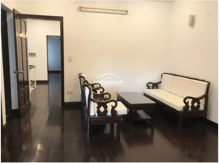 This living area features rich, dark-stained hardwood flooring that contrasts sharply with the minimalist white walls. The space is furnished with traditional dark wood seating featuring white cushions and flows directly into an adjacent room through a classic wooden door frame.