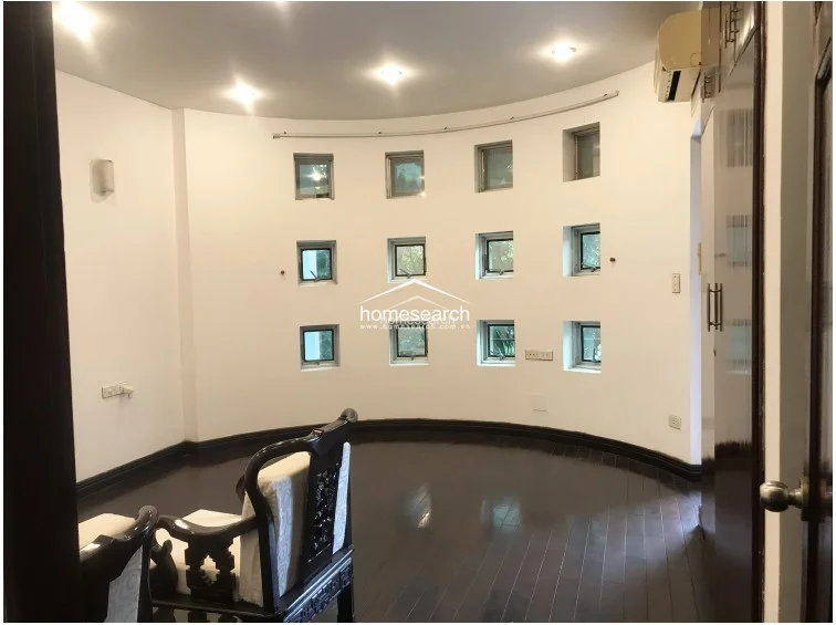 This unique circular room is defined by a series of small, square windows that provide consistent natural light and a modern architectural aesthetic. The space is finished with dark hardwood flooring, a wall-mounted air conditioning unit, and recessed ceiling lighting, maintaining a clean and well-maintained appearance.
