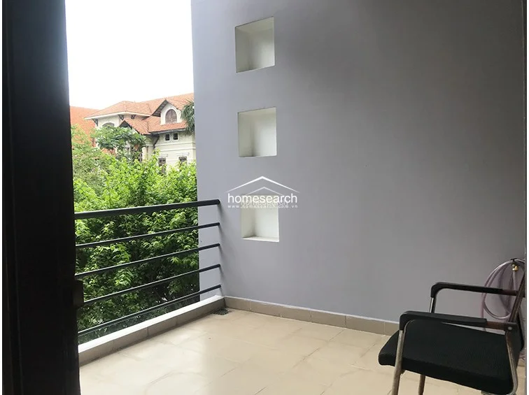 This outdoor balcony offers a private, serene space with a view of lush greenery and neighboring traditional architecture. It features a simple metal railing, tiled flooring, and built-in wall niches that provide a decorative touch to the neutral gray exterior wall.