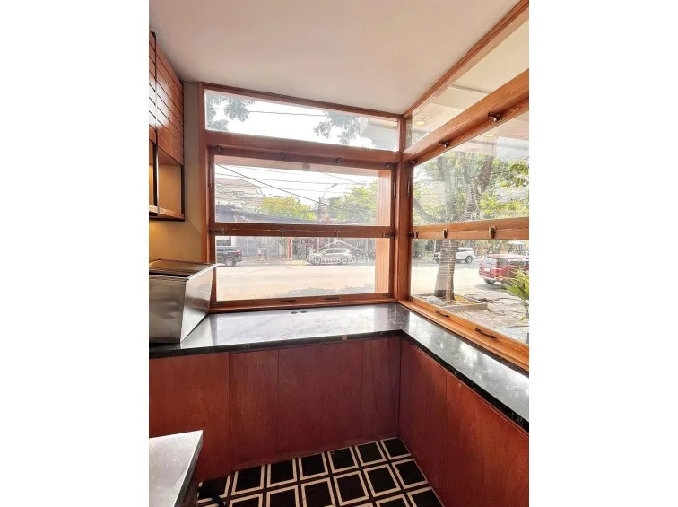 This kitchen corner features an L-shaped layout with dark granite countertops and warm-toned wooden cabinetry that provides ample storage. Large wooden-framed windows offer a clear view of the street, ensuring the workspace is bright and airy while maintaining a cohesive, high-quality design theme throughout the property.