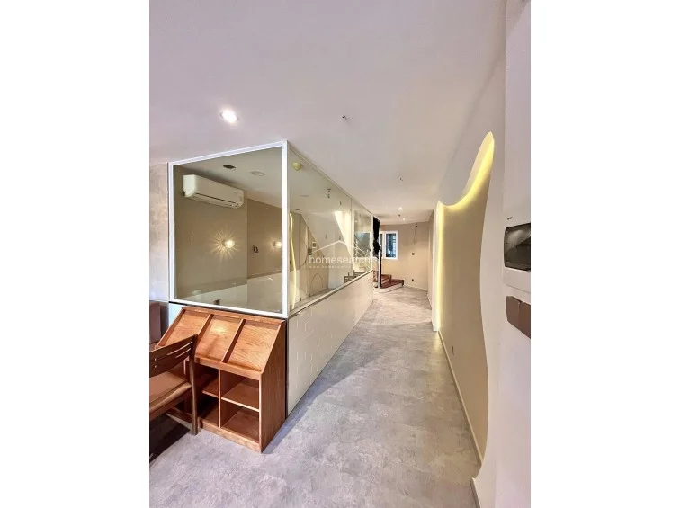 This interior space features a modern, open-plan layout with a unique glass-partitioned room that allows for visual connectivity throughout the area. The room is finished with light grey stone-look flooring and features a striking wall with built-in cove lighting, complemented by a wooden storage unit and a wall-mounted AC unit.