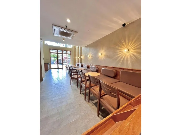 This commercial space features a long, linear layout with a dedicated dining area lined with built-in leather banquette seating and warm, accent wall lighting. The interior is finished with light grey stone-look flooring, a neutral beige wall palette, and a ceiling-mounted AC unit for climate control.