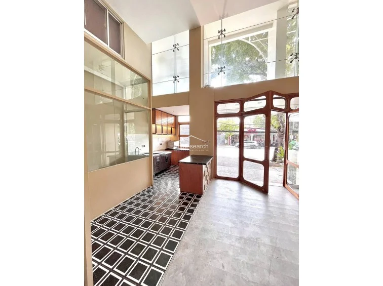 This wide-angle shot showcases the seamless flow between the front entrance, featuring large wooden-framed glass doors, and the rear kitchen area. The high-ceiling design and floor-to-ceiling glass windows flood the space with natural light, while the combination of wood cabinetry and stone countertops provides a polished, professional finish.