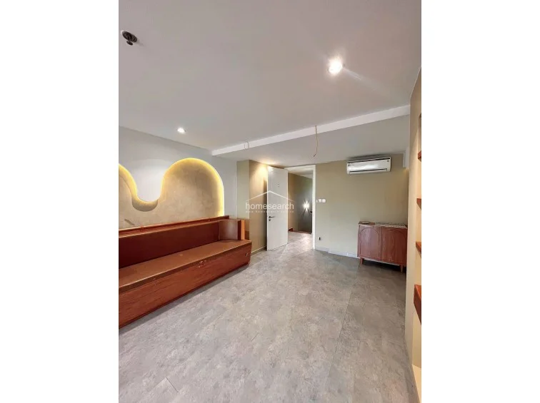 This living or lounge area is defined by a long, custom-built wooden bench seat set against a feature wall with illuminated architectural arches. The space maintains a minimalist aesthetic with neutral wall tones and consistent grey flooring, while a wall-mounted AC unit and recessed ceiling lights provide functional comfort.