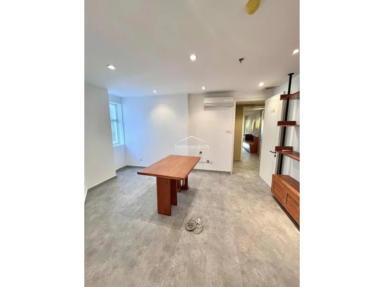 This versatile room is configured as a workspace, featuring a central wooden desk and an open-shelving unit that adds both storage and character. The room is brightly lit by a side window and recessed ceiling lights, with a wall-mounted AC unit ensuring climate control in this clean, modern environment.