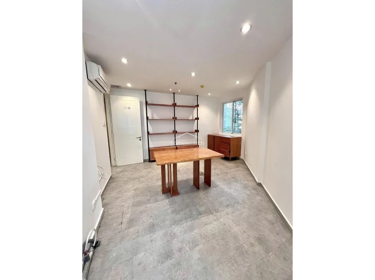 This versatile room features neutral white walls, grey tiled flooring, and recessed ceiling lighting, creating a clean and modern aesthetic. It is furnished with a minimalist wooden table and a floor-to-ceiling open shelving unit, while a wall-mounted AC unit and a window providing natural light complete the functional space.